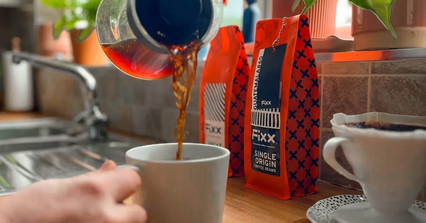 يد تصبّ قهوة مقطّرة في كوب على طاولة المطبخ، بجانب كيس FiXX Guatemala Single Origin وقمع تقطير