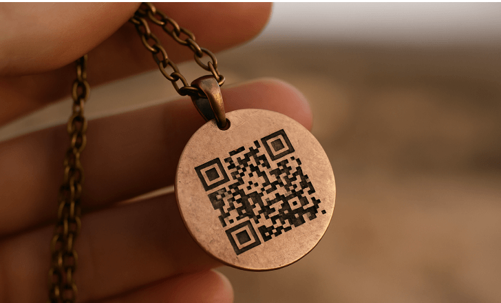 A hand holding a copper pendant necklace engraved with a QR code, against a blurred outdoor background.
